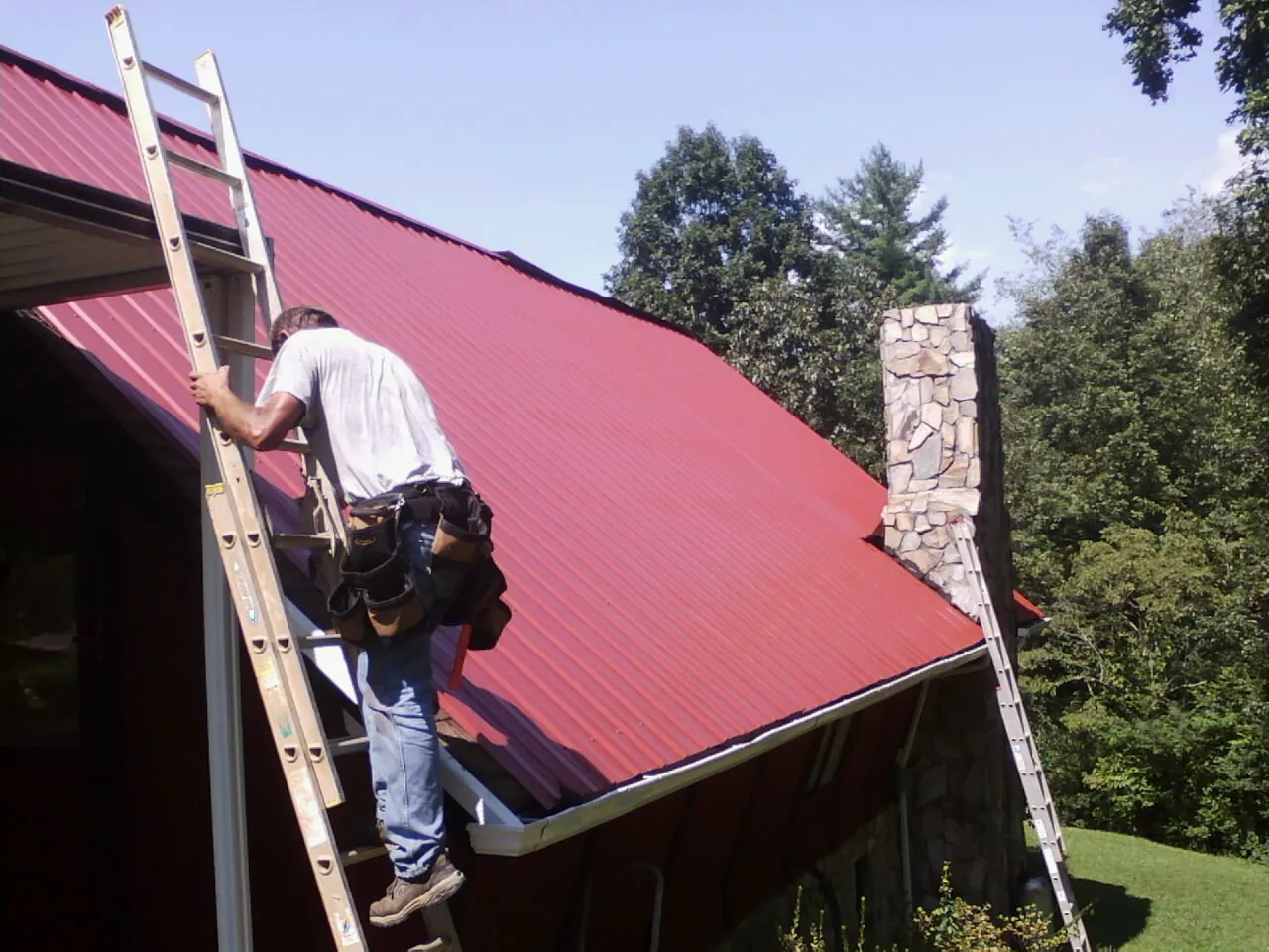 Roofer on a ladder beside a red metal roof and stone chimney — Soffit & Fascia Repair by Roofing Brothers in Dickerson Run